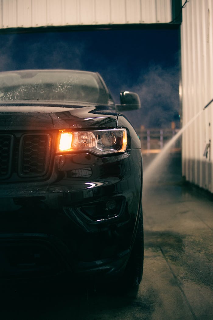 A sleek black car undergoing a wash at night, highlighting the headlight and water reflections.
